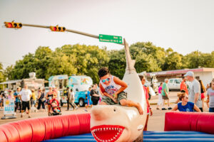 Child riding a shark-shaped inflatable ride amid a crowded outdoor festival scene.