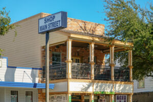 La Porte Main Street 2 Thomas Park - Texas Hotel and Lodging Brick storefront with a tall pole-mounted sign reading 'SHOP MAIN STREET' against a clear blue sky.