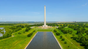 La Porte San Jacinto Monument Terry Thorn - Texas Hotel and Lodging Tall white obelisk with a star on top in a grassy park, beside a long reflecting pool under a blue sky.