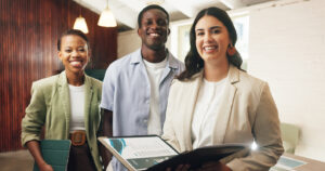 Three colleagues stand together in a bright office, smiling at the camera, with folders and a binder in hand.