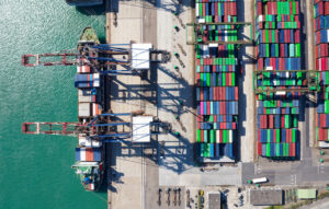 Aerial view of a container port with colorful stacked shipping containers and large cranes over turquoise water.