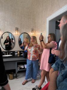 Driskill hotel tour_ FCSTAT - Texas Hotel and Lodging People in a stylish hotel restroom, a woman in floral top and blue pants stands by the counter while others pose and take photos in front of round mirrors.