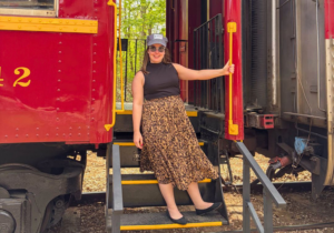 Smiling woman in a black sleeveless top and leopard print skirt posing on the steps between two red train cars, gripping a yellow handrail as she poses outdoors in sunlight.