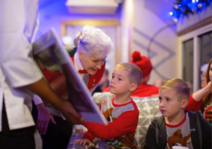 Visit Palestine_11 - Texas Hotel and Lodging An elderly woman with white hair hands a card or drawing to a young boy at a festive indoor gathering with blue lighting and holiday decor in the background.