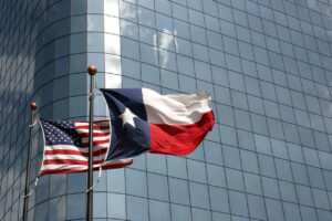 American flag and Texas state flag fluttering on flagpoles in front of a glass office building